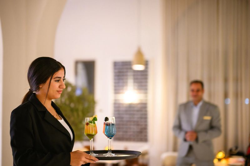 Arab waitress skillfully carrying a tray with colorful drinks in a modern hotel restaurant. The lighting is warm and inviting, reflecting off the sleek decor. A male guest in a suit stands in the background, creating a sophisticated dining atmosphere.