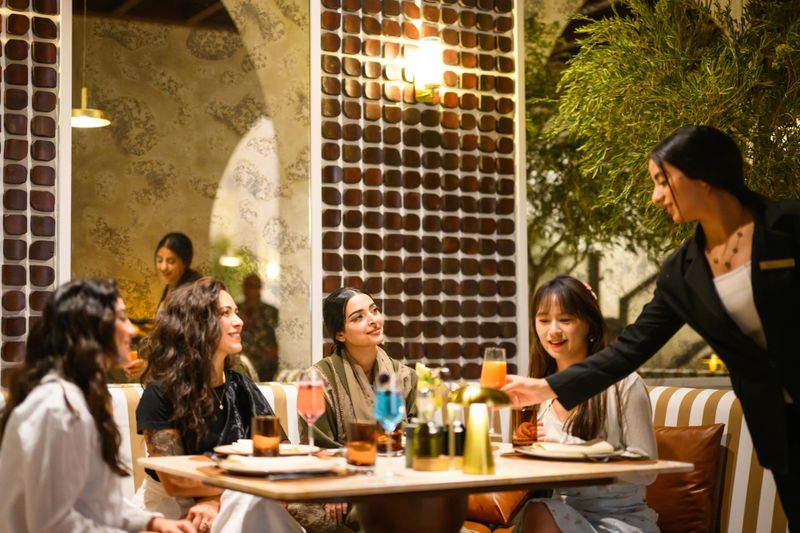Young Arab waitress serving a refreshing beverage to a diverse group of happy women dining together inside an elegant luxury hotel restaurant. The setting features decorative wooden screens and textured walls, utilizing rich, warm ambient lighting that creates an intimate evening atmosphere. Female guests engaging in relaxed conversation while enjoying the upscale dining experience.
