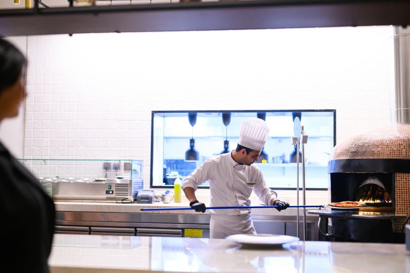 Arab male chef wearing a white toque and uniform using a long metal peel to handle flat bread inside a traditional wood-fired oven. An Arab female waitress waits nearby at the stainless steel kitchen pass counter, ready for service. The bright, modern commercial kitchen features white tiled walls and professional equipment, emphasizing efficiency and high-end dining preparation.