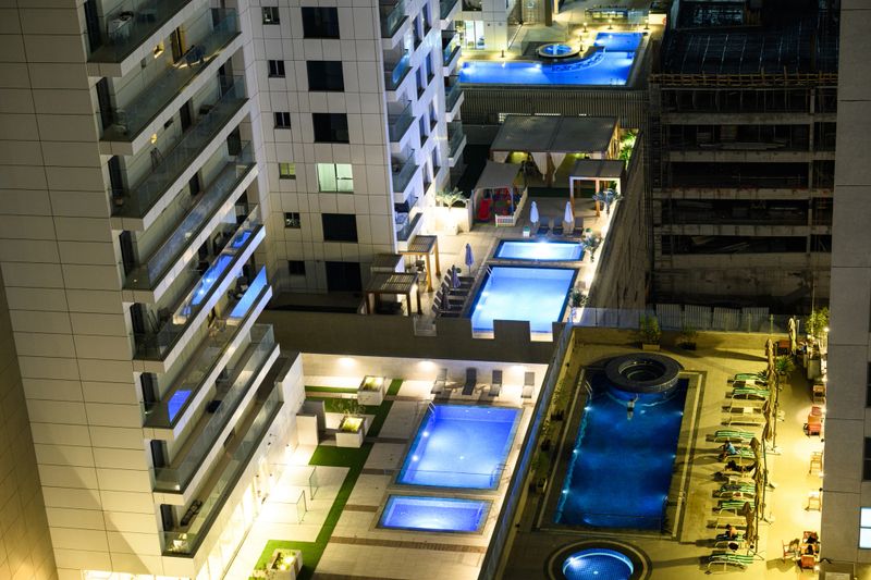 Elevated view of multiple illuminated rooftop swimming pools surrounded by modern apartment buildings at a luxury hotel in Dubai, United Arab Emirates. The pools are lit with colorful underwater lighting, creating a vibrant atmosphere against the night sky. The hotel's architecture features glass and steel elements.
