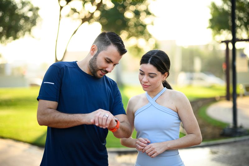 A mid-adult Caucasian couple checking their fitness watch together after an early morning exercise session in a lush, green park. The pair appear focused and engaged as they review their workout data, surrounded by the tranquil park setting.