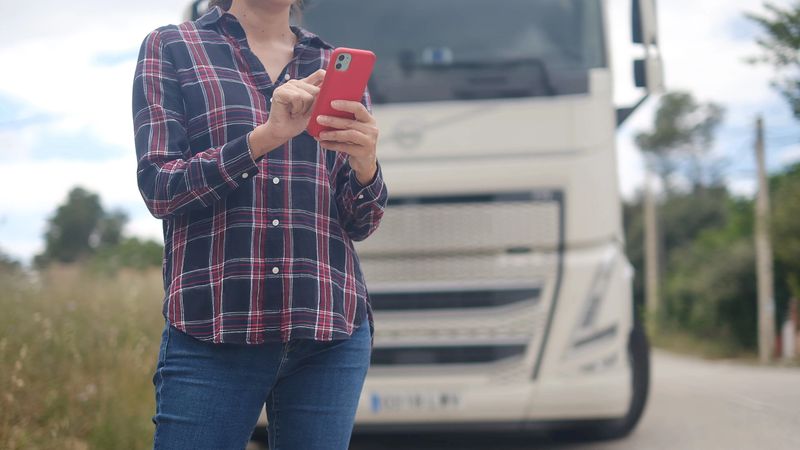 Woman standing next to a truck, using a smartphone to manage logistics, tracking routes, and communicating for efficient freight transportation services