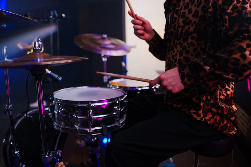 A musician is playing the drums in a studio setting. The drummer uses sticks to hit the snare and cymbals while the environment shows bright lights.