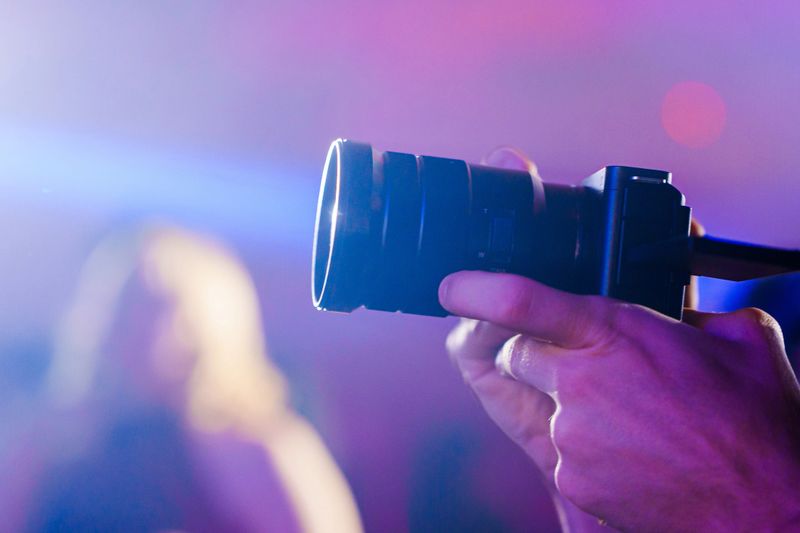 A person holds a camera in one hand and points it towards the scene at a lively event with colorful lights and people in the background, capturing moments.