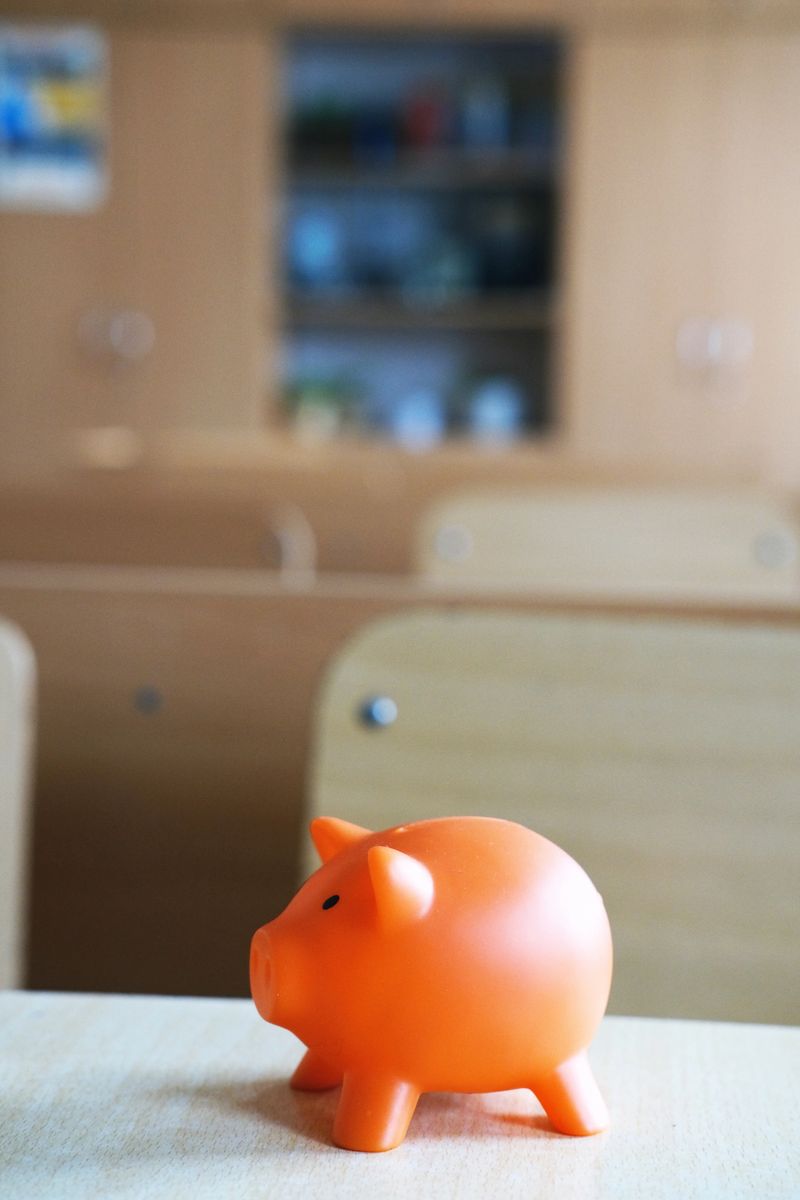 Bright orange piggy bank sits on a wooden desk in a school classroom, representing financial planning and school expenses, with a blurred background of classroom elements