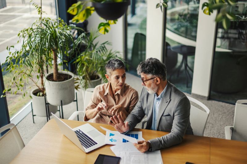 Mid-adult colleagues review financial reports and use laptop, tablet and desktop in a modern, plant-filled office, focused on strategy, collaboration and eco-friendly workplace productivity