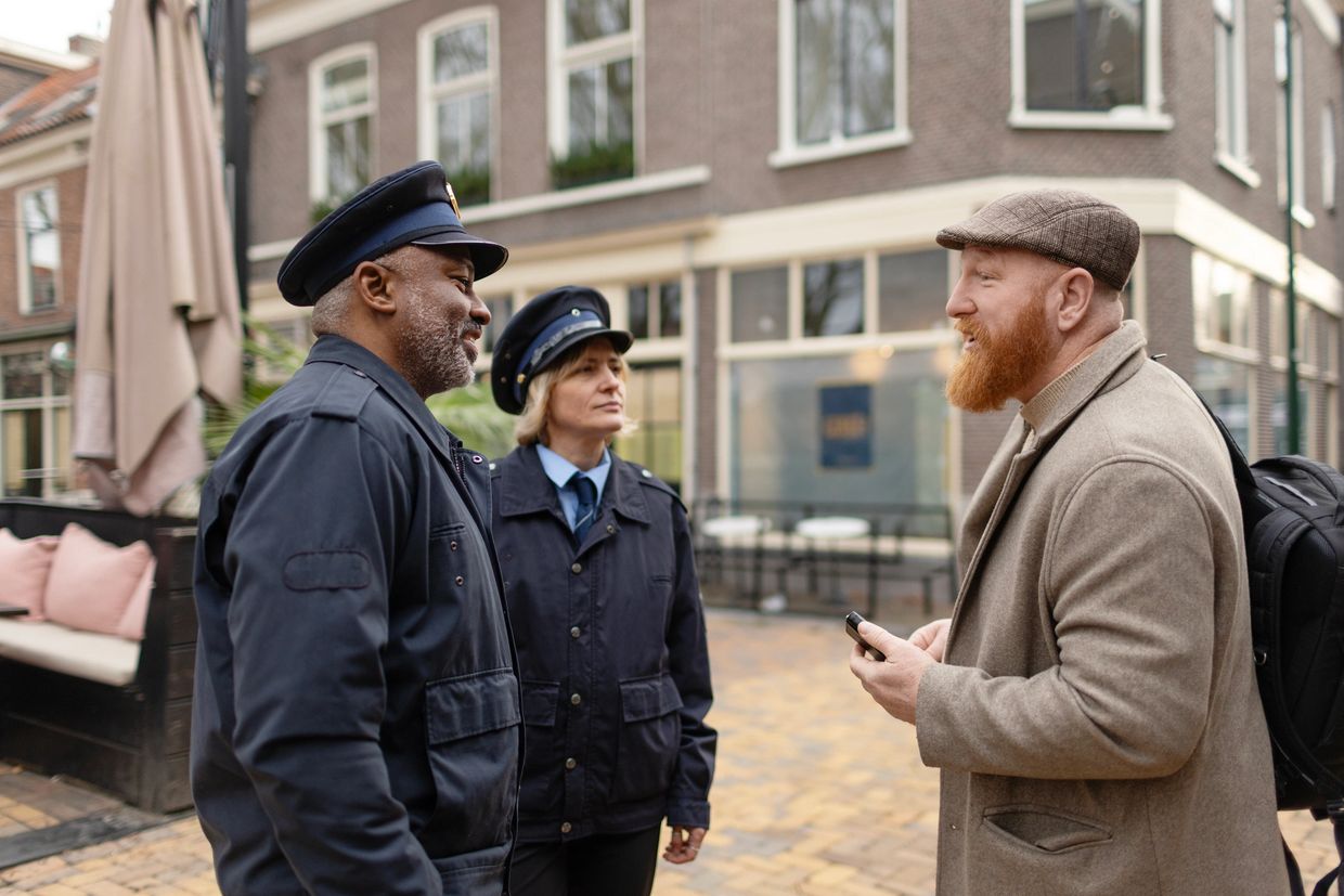 Two police officers talking to a man with a beard and flat cap on a city street.