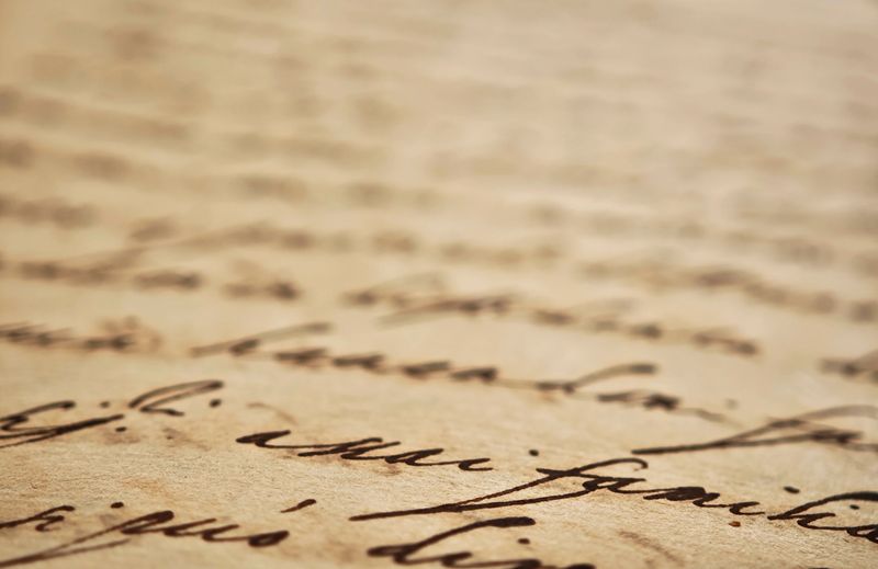 Extreme closeup of old handwritten manuscript on yellowed paper with cursive ink writing, shallow depth of field soft blurred background, vintage document texture history, literature, poetry, archival
