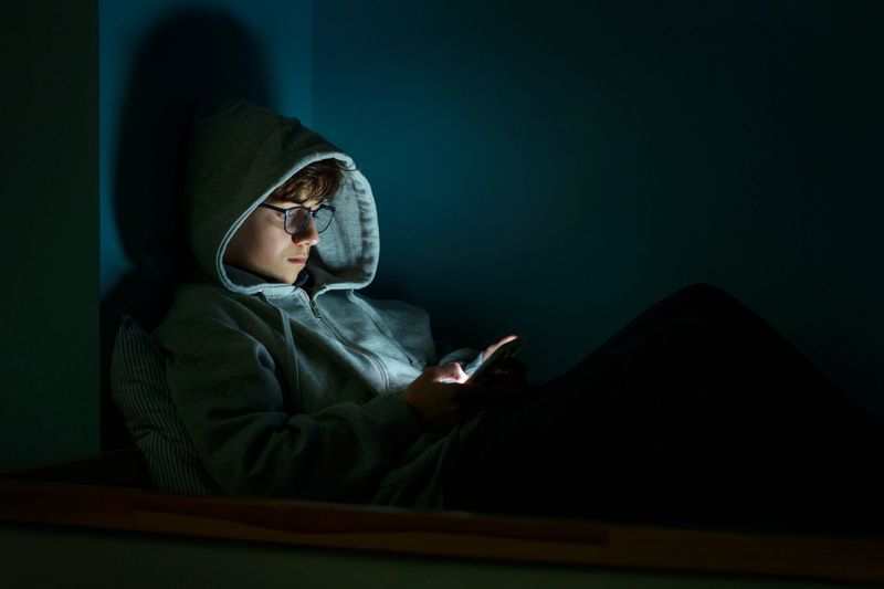 Teenager wearing a hoodie and glasses is using a glowing smartphone in a dark room
