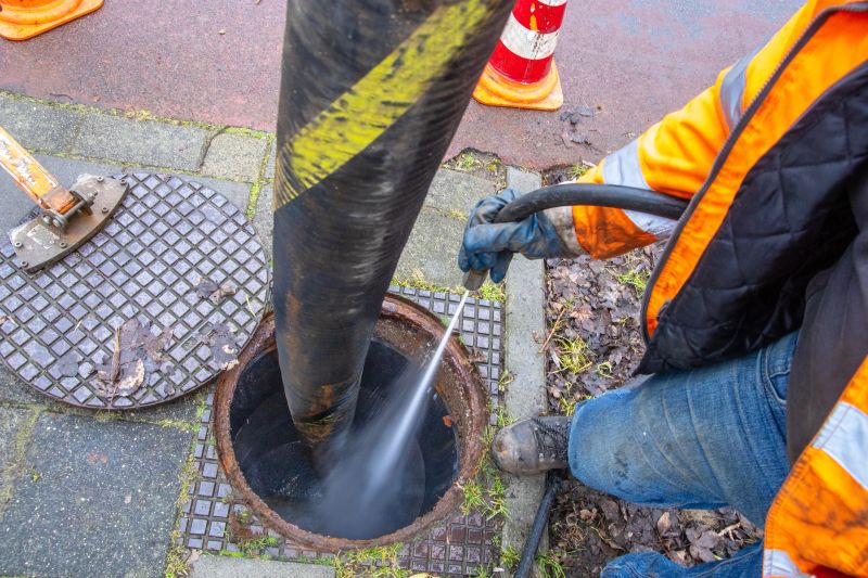Workers clean a sewer manhole using a high-pressure water hose and vacuum system from a sewer truck. After cleaning, a sewer inspection camera is lowered into the manhole to document the condition of the underground infrastructure. Urban sewer maintenance in a public area.