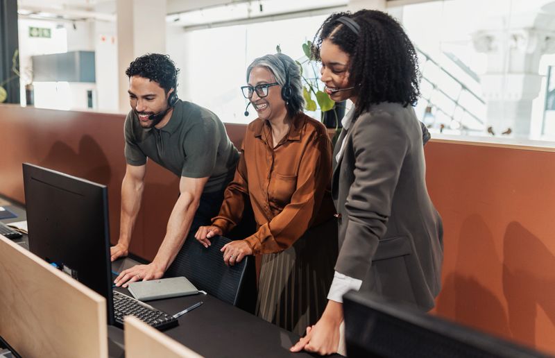 Young customer support operators wearing headsets, standing with female supervisor, all smiling happily and learning from work tutorial on desktop computer in call center