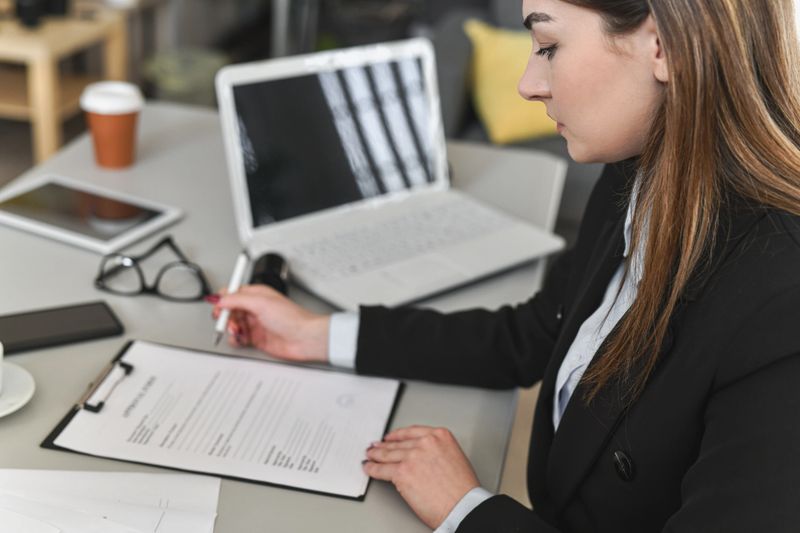 A focused professional woman signs a contract at a clean office desk, surrounded by a laptop, tablet, clipboard, and documents. The scene conveys business, professionalism, and careful review of paperwork.