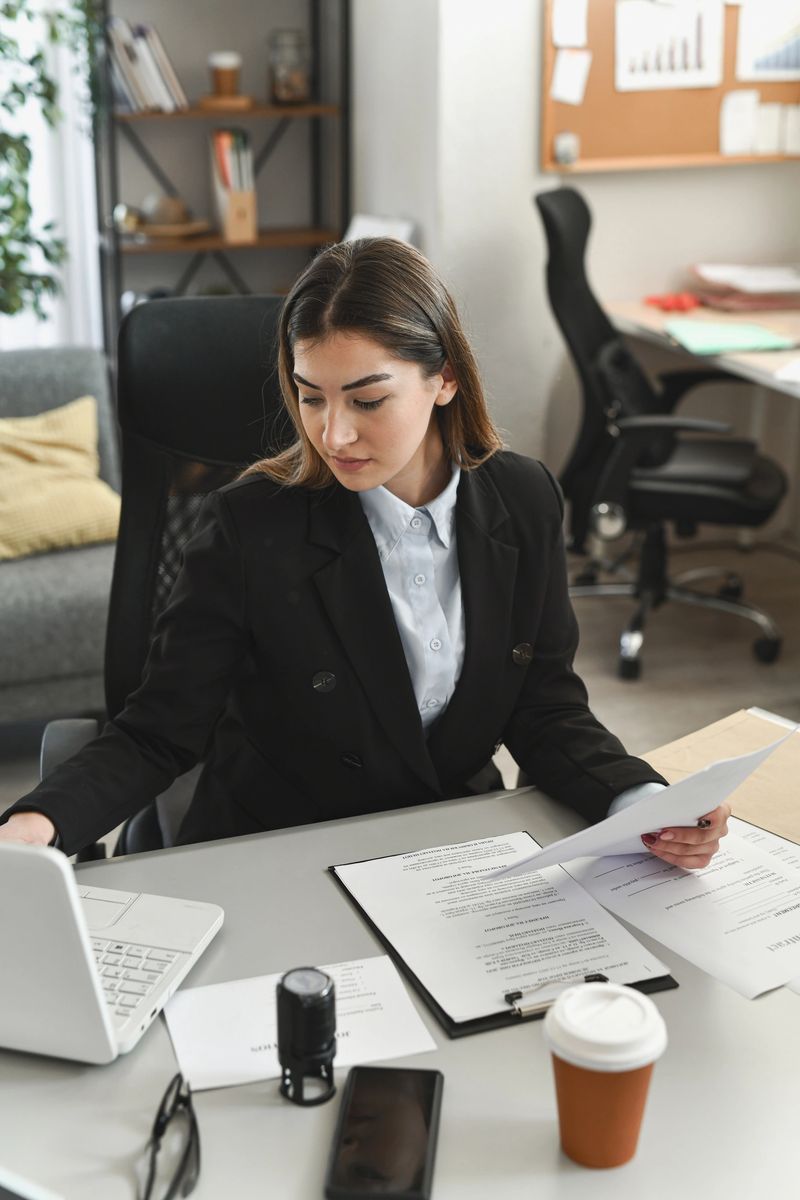 A focused businesswoman sits at a clean office desk, reviewing papers while a laptop and coffee are at hand, conveying productivity, organization, and professional focus in a modern workspace.