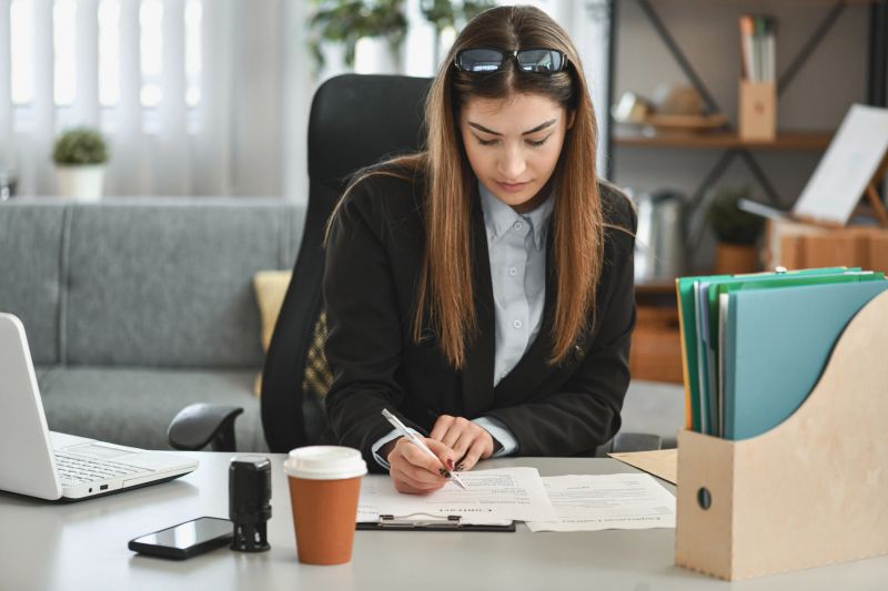 A focused professional woman signs documents at a clean office desk, surrounded by a laptop, phone, coffee, and organized folders. This image conveys business, efficiency, and careful paperwork.
