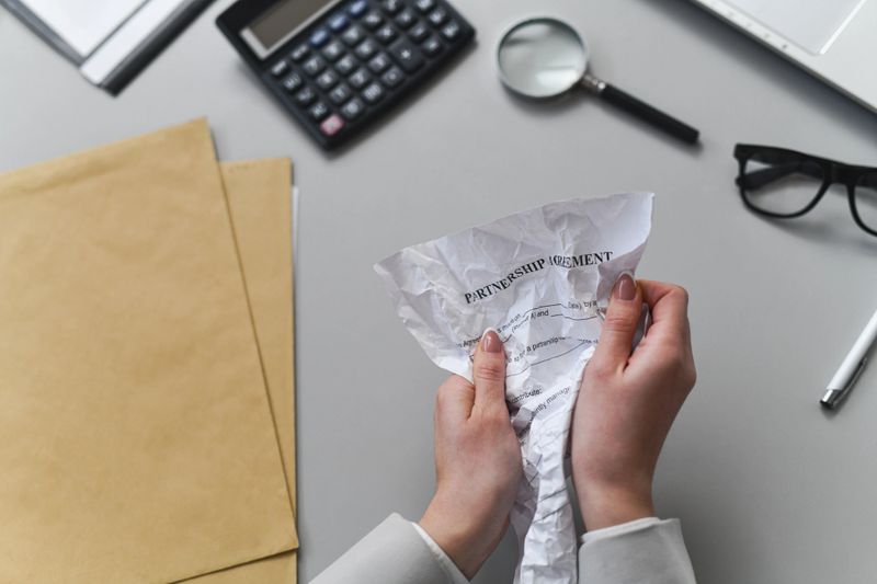 Hands crumple a document labeled 'Partnership Agreement' in a professional office setting with a calculator, magnifier, and folders in the background. The moment conveys stress, negotiation, and paperwork.