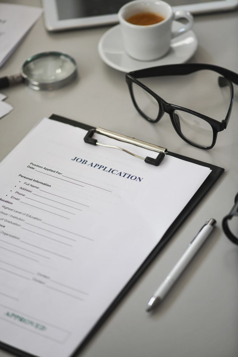 A focused businesswoman signs and reviews a job application form clipped to a clipboard in a sleek office, with glasses in the background, conveying professionalism and recruitment processes.