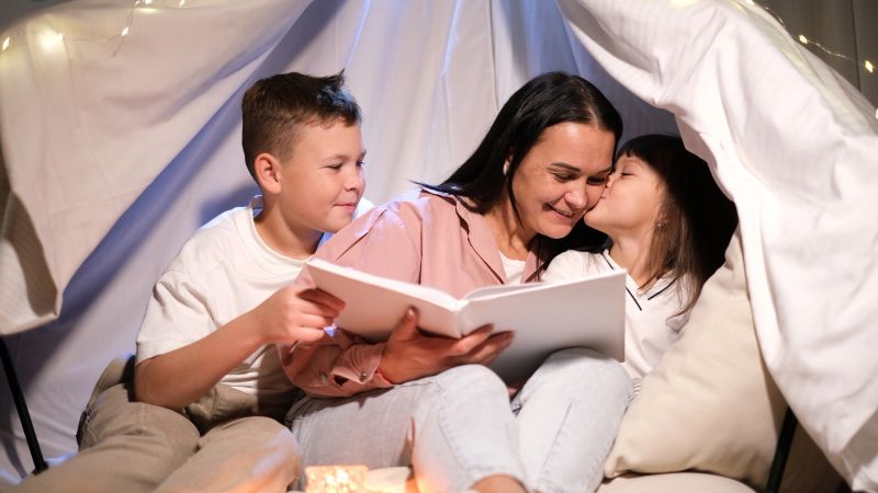 Premium lifestyle footage capturing childhood magic and nurturing family bonds A mother creates a memorable experience by reading a story to her two young children inside a whimsical blanket fort during a cozy evening This scene illustrates concepts of imaginative play quality family time educational engagement and creating special rituals at home Ideal for advertising children's books family focused brands home furnishings or editorial content on modern parenting and creative childhood development