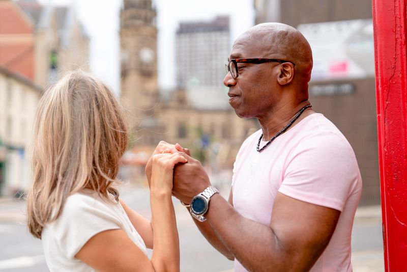 A man and a woman stand together on a busy city street. They hold hands and look at each other with smiles. Tall buildings surround them in the background.