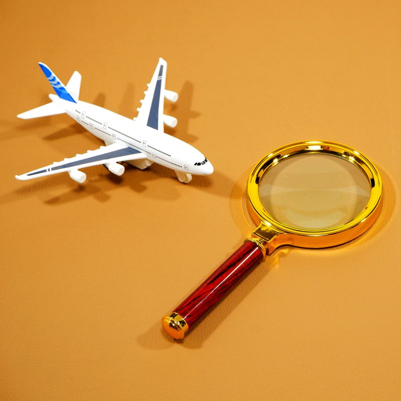 Classic handheld magnifying glass with a golden rim and wooden handle positioned next to a detailed miniature white passenger jet model. This juxtaposition suggests close examination or careful planning required for global aviation, travel logistics, or air transport auditing processes.