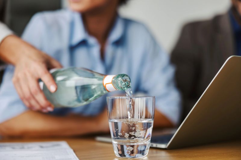 Woman's hand pouring refreshing water from a glass bottle into a drinking glass on a wooden table, emphasizing healthy hydration during a busy workday or business meeting