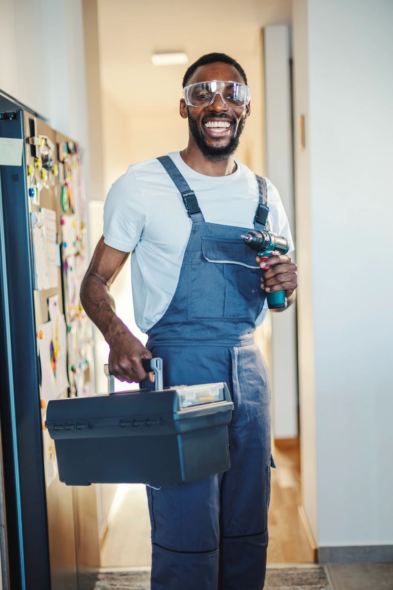 African american male handyman smiling confidently, wearing work overalls and safety goggles, holding a toolbox and a power drill, providing reliable home repair and improvement service