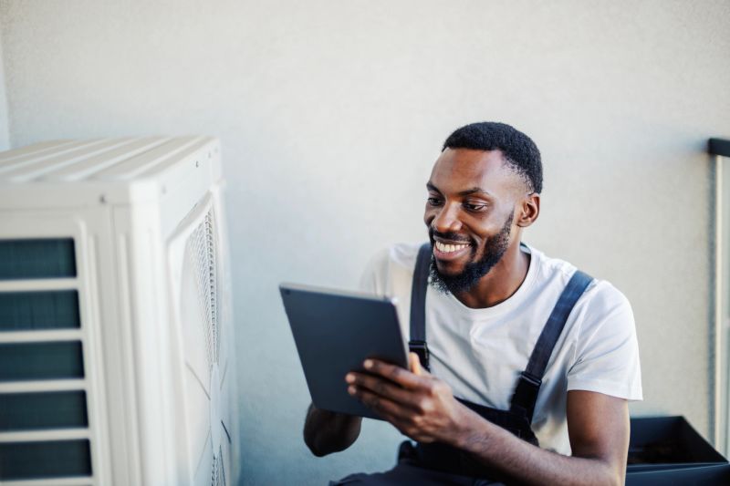 African american male hvac technician smiling while holding a digital tablet and inspecting an outdoor residential air conditioning unit, performing maintenance and service