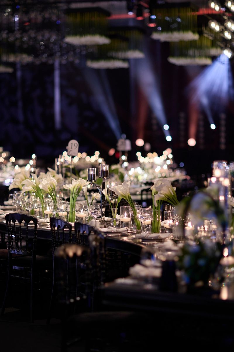 Elegant dining table set for a luxury event, featuring white floral arrangements, refined tableware, and soft candlelight creating a warm and sophisticated atmosphere. The shallow depth of field and glowing bokeh lights enhance the sense of intimacy and exclusivity, making this image ideal for concepts related to high-end weddings, formal receptions, fine dining experiences, and premium event styling.