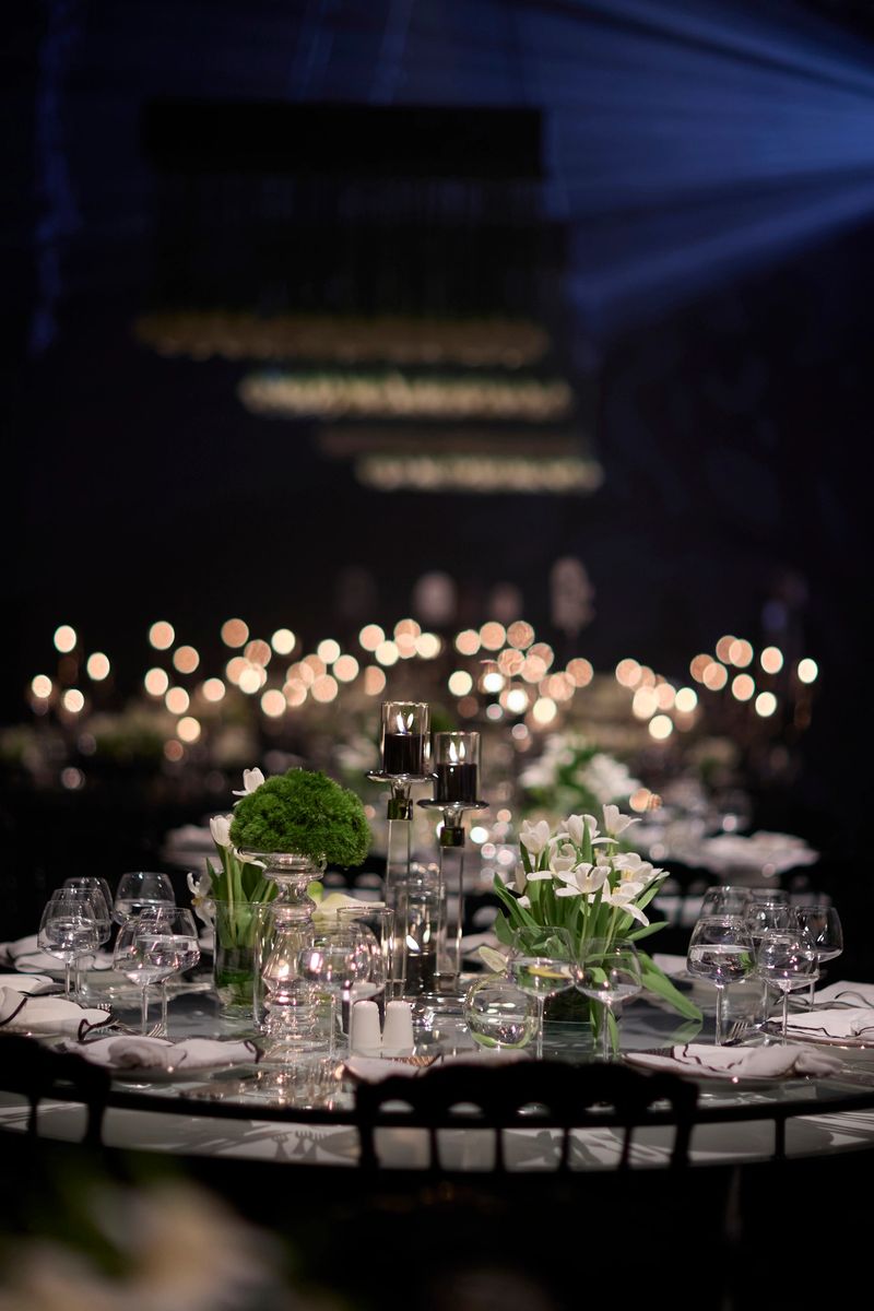 Luxury event table decor featuring elegant candlelit settings and suspended floral installations above the tables. The refined composition highlights modern event styling, soft ambient lighting, and a sophisticated atmosphere, ideal for concepts related to high-end weddings, exclusive celebrations, fine dining experiences, and premium hospitality design. The dark background enhances depth, contrast, and a sense of intimacy. stock photo