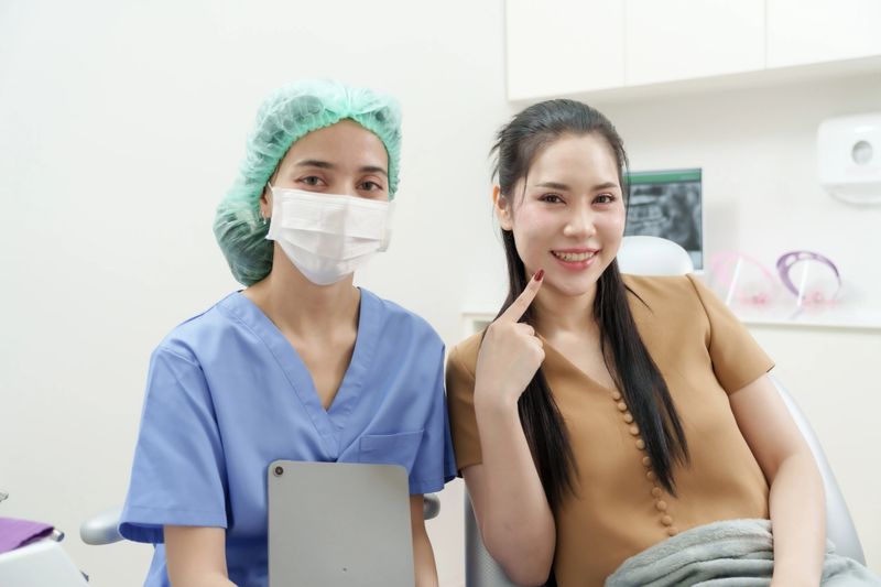 Asian woman smiling next to female dental assistant in uniform and mask inside clinic. Patient pointing to teeth after receiving instructions on dental care routine following braces treatment.