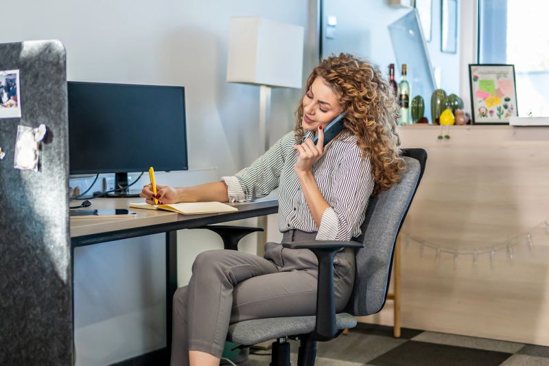 Young adult woman busy working in an office, communicating on the phone while taking notes in her notebook