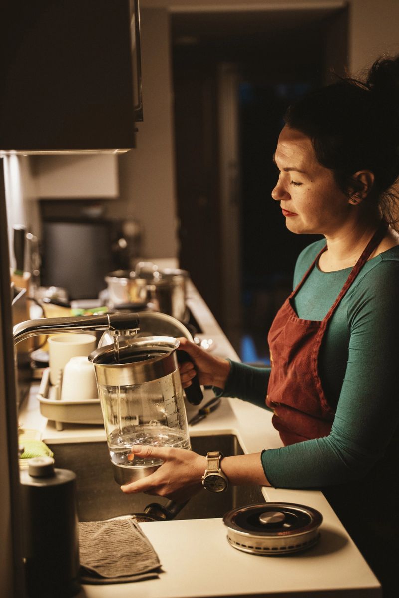 Woman in kitchen fills a glass kettle with purified water from a home water purifier. She appears focused and content, ensuring her water is clean and safe for use.