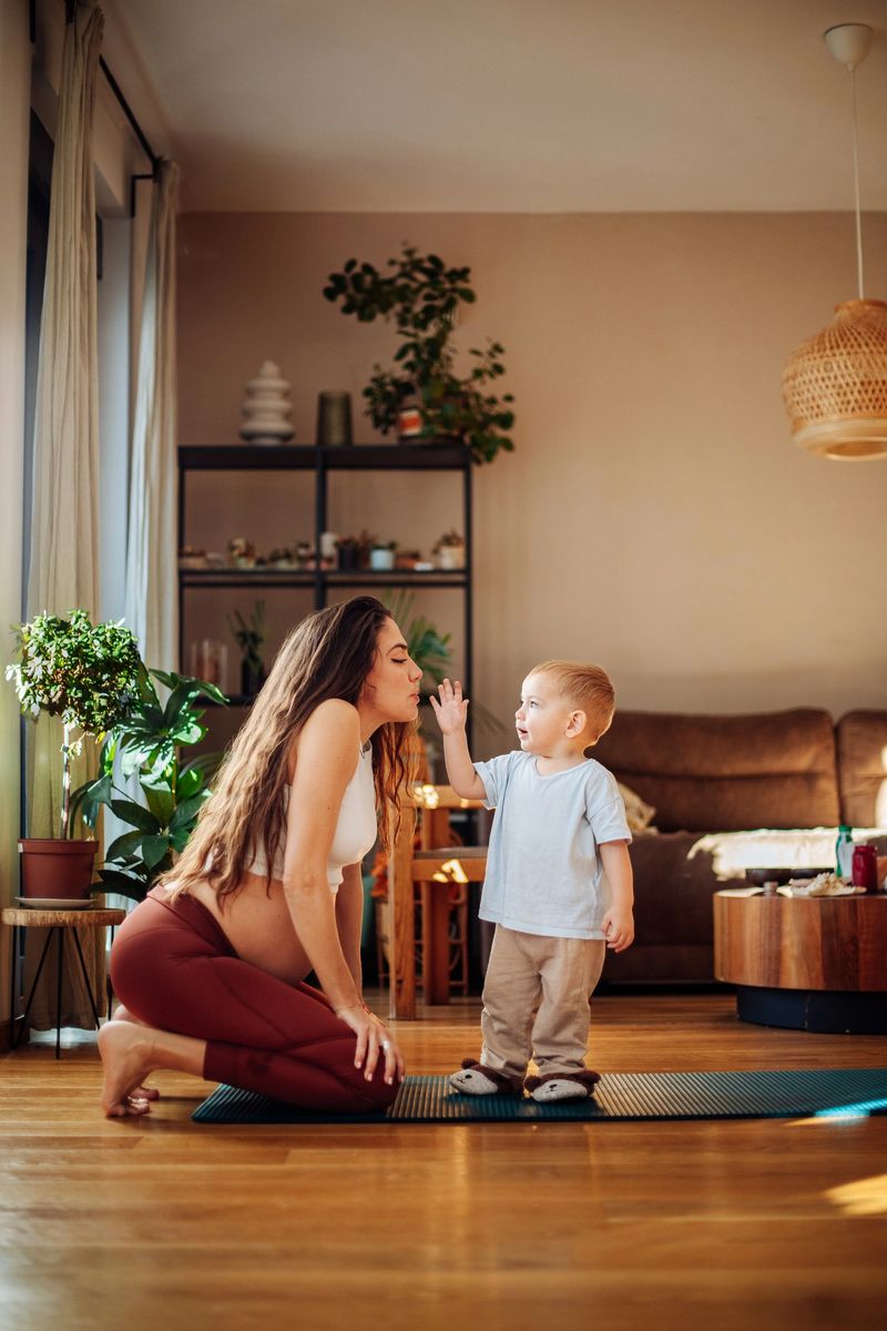 A pregnant woman in athletic wear kneels on a yoga mat, interacting playfully with her young son who stands before her, both smiling in a sunlit home environment
