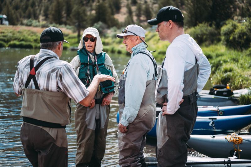 Middle-aged and senior men stand in shallow water discussing plans beside inflatable rafts before starting a fly fishing trip in Central Oregon.