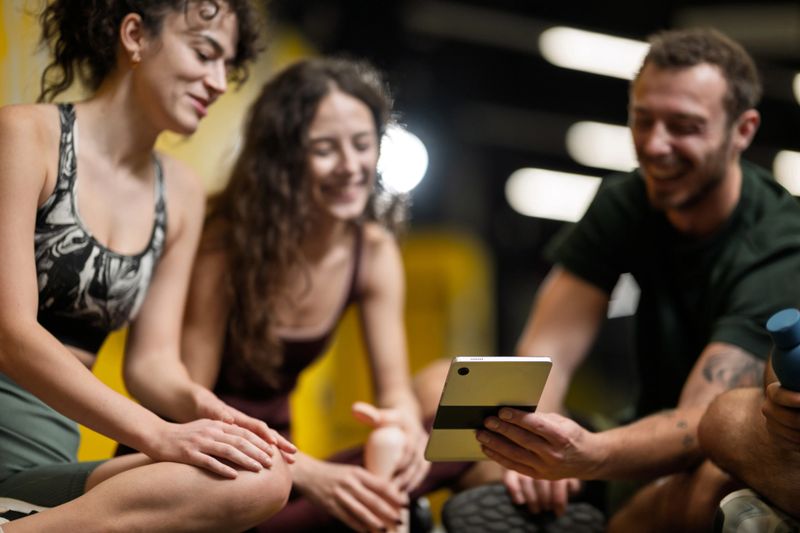 Three friends gather on the gym floor to share fitness tips. They look at a tablet together while enjoying their workout time in the fitness studio.