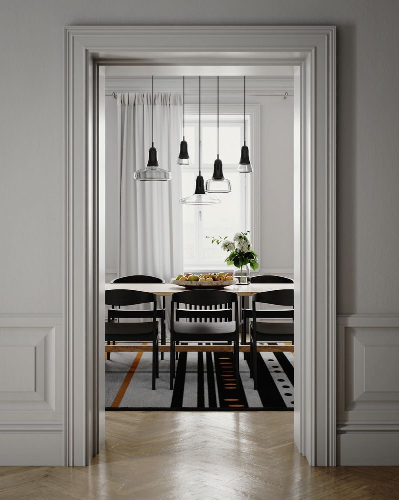 Digitally generated view of an elegant modern dining room seen through a classic white doorway in a bright apartment. The wooden table with black chairs, pendant lights and fruit bowl sits on a striped rug, framed by crisp wall moldings and parquet floors. Minimalist Scandinavian interior architecture concept with strong framing and copy space.