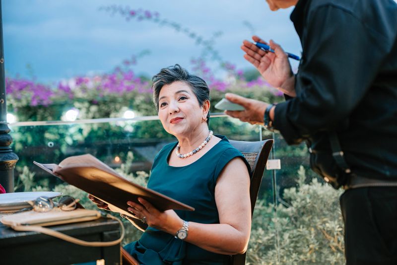 sophisticated senior Asian woman with short grey hair sits at a dinner table holding a menu. She looks up with a pleasant smile at a waiter who is taking her order. The background features soft twilight bokeh and purple flowers.
