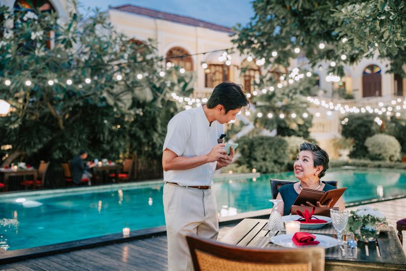 Senior Asian woman with grey hair sits at an elegantly set table by a turquoise swimming pool at dusk. She holds a menu and looks up to talk with a young male Asian waiter wearing a white shirt and tan trousers. Warm string lights decorate the background.