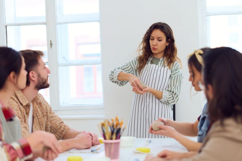 A bright, airy studio as a teacher in a striped apron demonstrates a craft technique to a group of friends. They sit around a table, hands-on, focused and collaborative, enjoying a creative workshop.