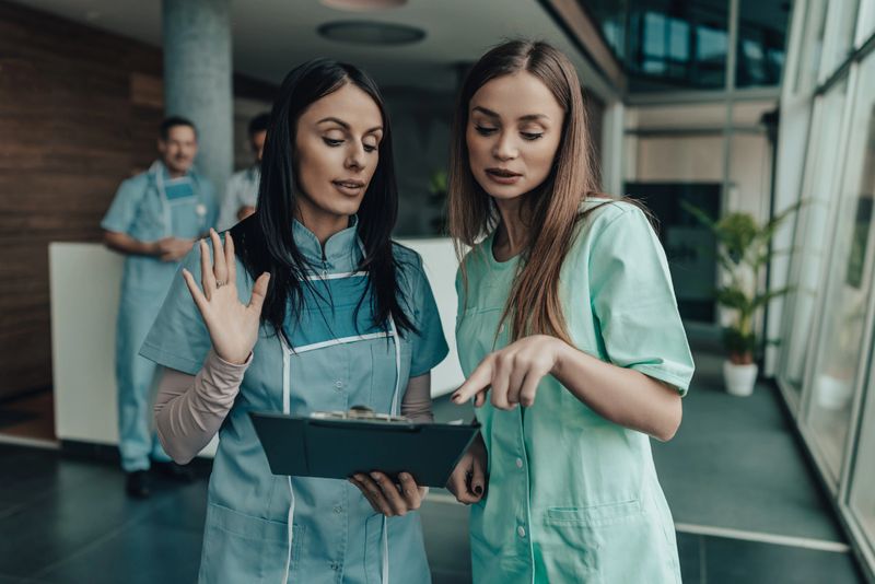 Two healthcare professionals in scrubs review a clipboard while one points, highlighting teamwork, guidance, and learning in a contemporary clinic setting.