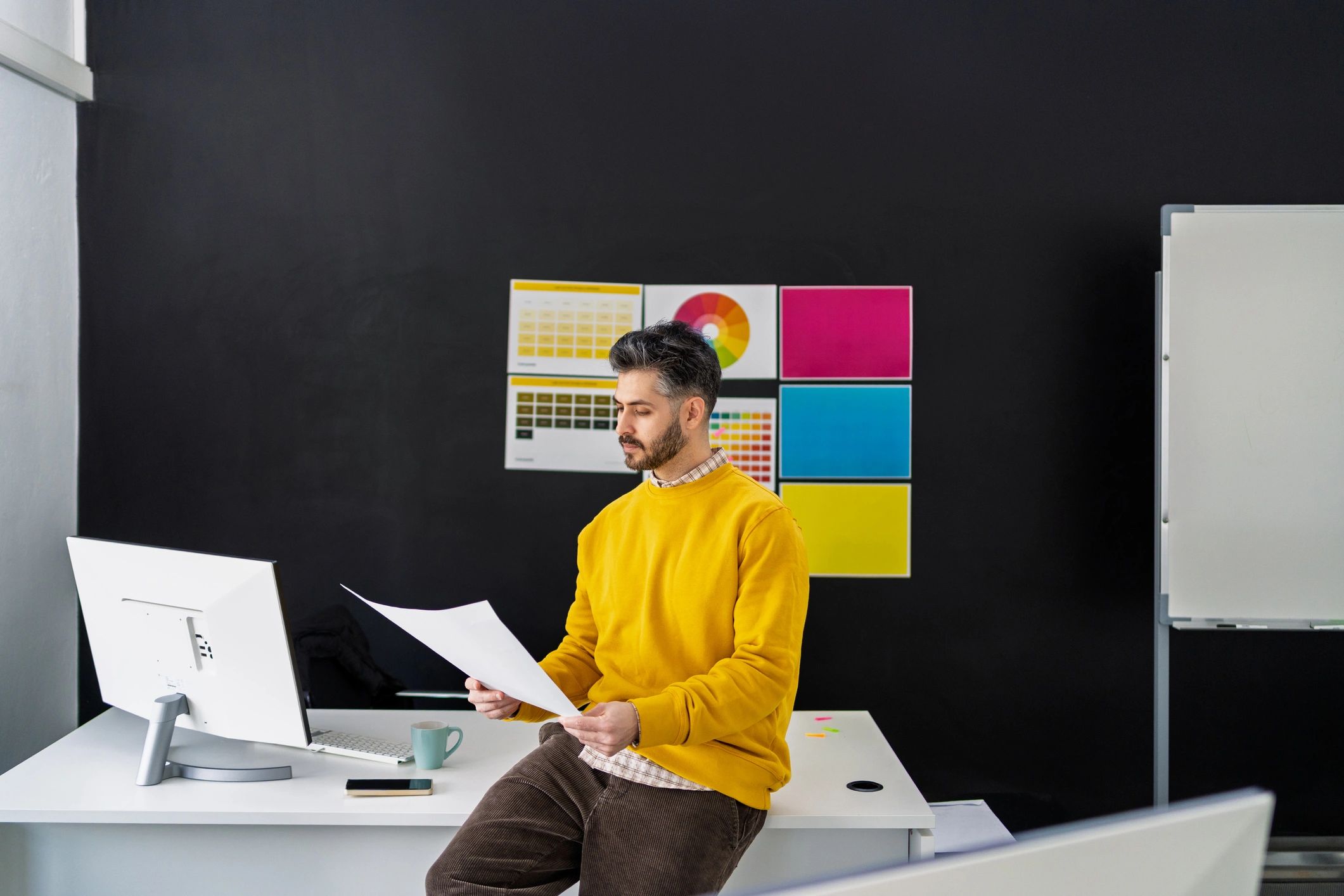 Man in yellow sweater sitting on a desk reading a paper.