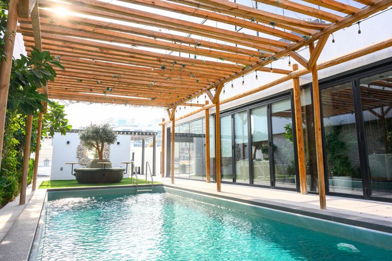 Contemporary rooftop swimming pool featuring a wooden pergola, string lights, and modern architecture, providing a luxurious outdoor living space with sunlit tranquility