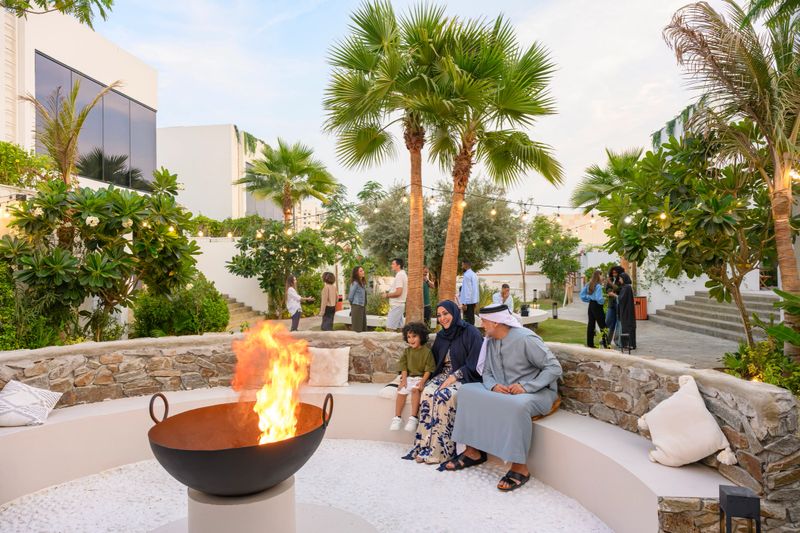 Family members and several guests are relaxing around an outdoor fire pit in a modern luxious garden setting, socializing and enjoying the warm evening outdoors