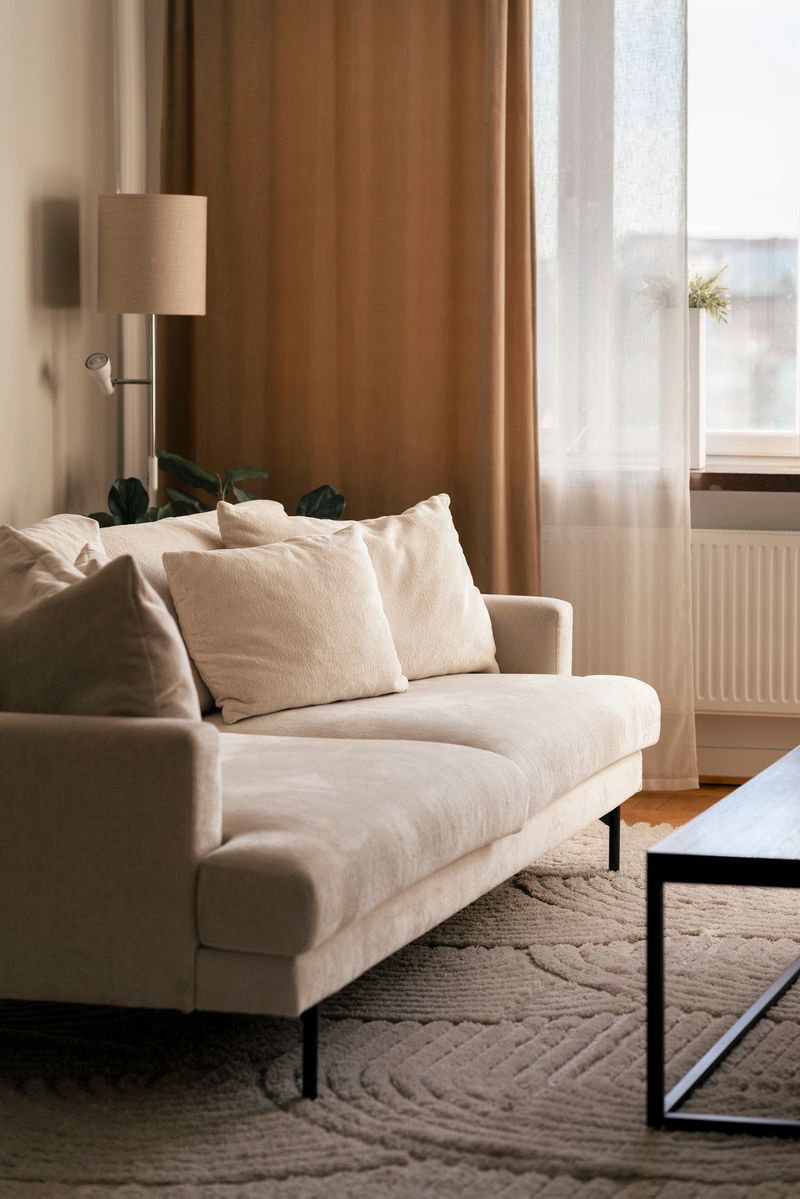 A warm, neutral living room scene featuring a plush beige sofa, soft natural light from a large window, a floor lamp, and leafy plants. Calm, inviting space perfect for relaxation and home comfort.