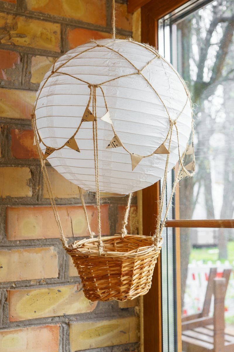 Decorative handmade paper hot air balloon lamp with wicker basket hanging indoors near window. Cozy creative interior decor concept with natural light and warm textures.
