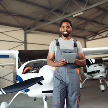 Smiling aircraft mechanic holding a tablet in front of a small airplane in a hangar.