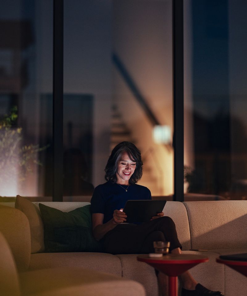 A senior woman sits comfortably on a sofa at night, smiling while reading or working on a tablet. Warm ambient lighting creates a cozy, relaxed home office mood.