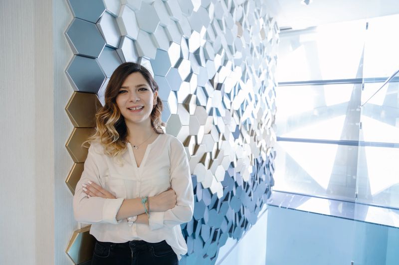 Confident young professional businesswoman wearing white blouse standing with crossed arms and looking at camera in bright modern office corridor with abstract geometric wall pattern, corporate lifestyle, leadership concept, stylish workplace environment, natural light, copy space