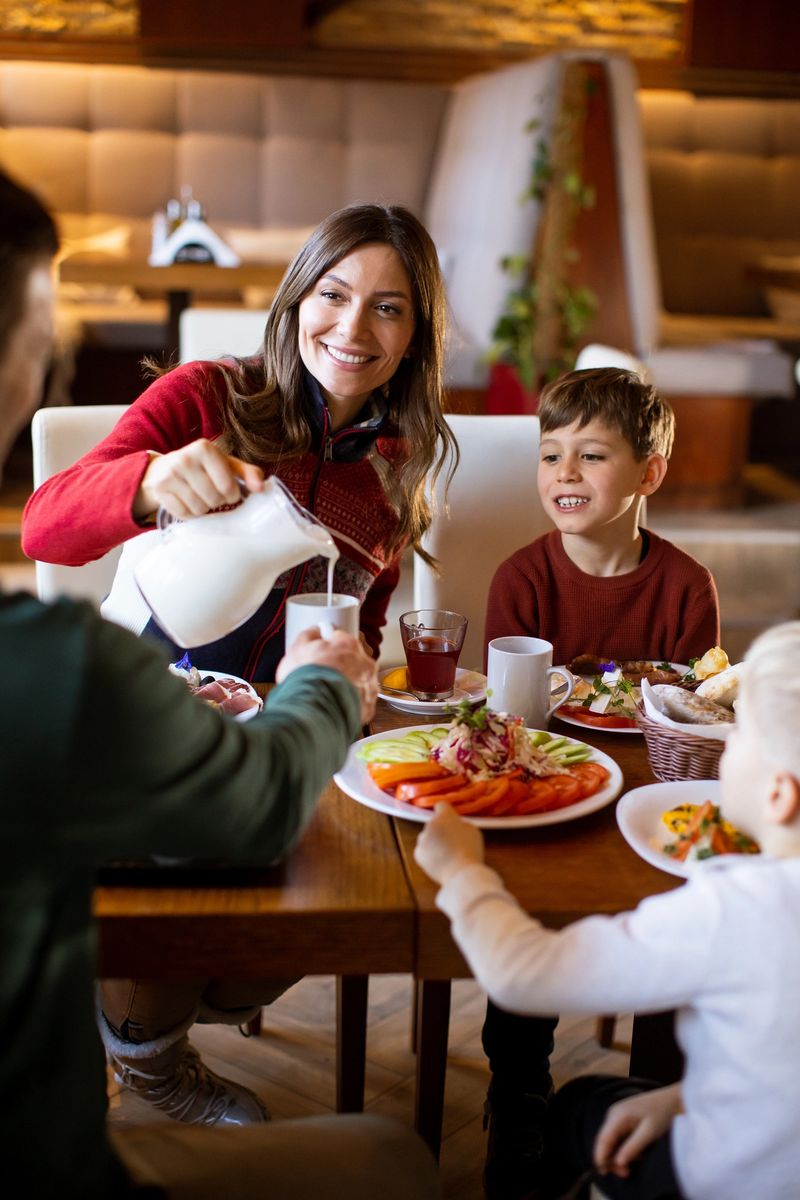 A woman pours milk for her son while a young boy watches at a cafe. The table is filled with plates of food and drinks. They all smile and enjoy the morning together.