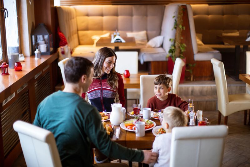 A family sits around a table sharing breakfast. They enjoy food and drinks while chatting and laughing. The restaurant has a warm atmosphere and comfortable seating.