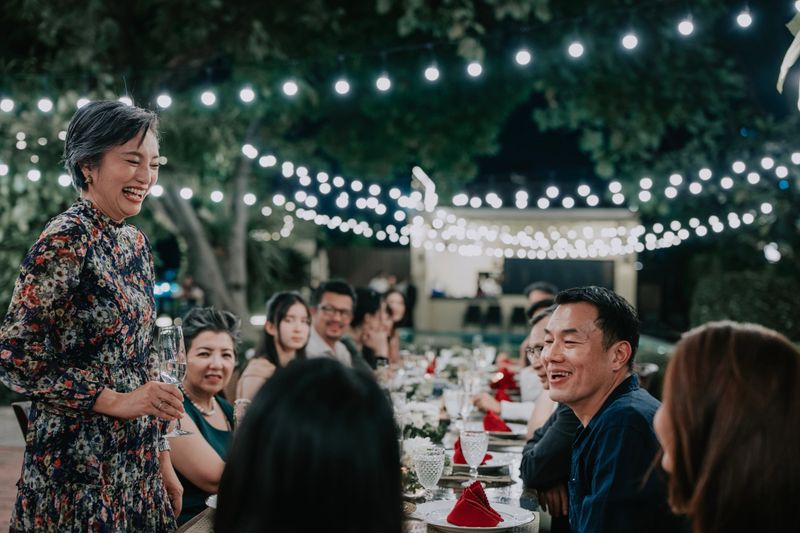 A joyful mature Asian female stands with a glass of sparkling wine while giving a speech at an outdoor night dinner with Asian friends and family.